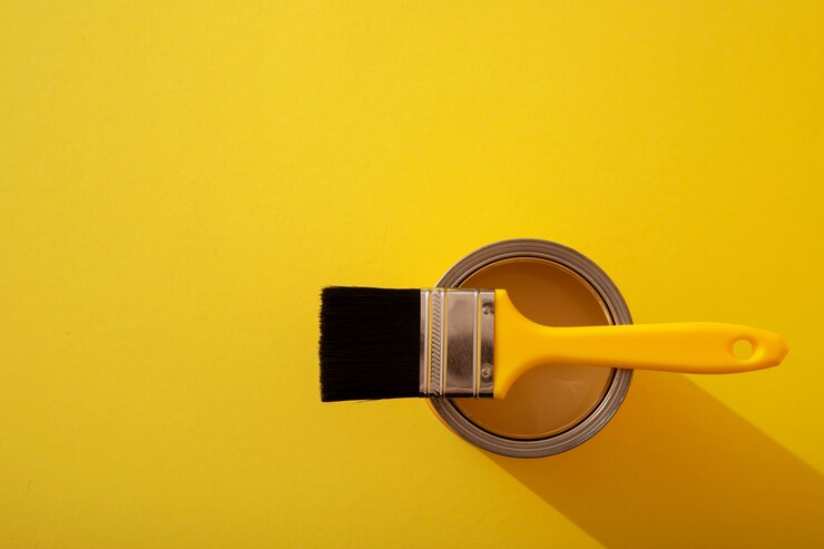 Top view of a paintbrush resting on an open can of paint against a bright yellow background, symbolizing painting, coatings, and DIY projects