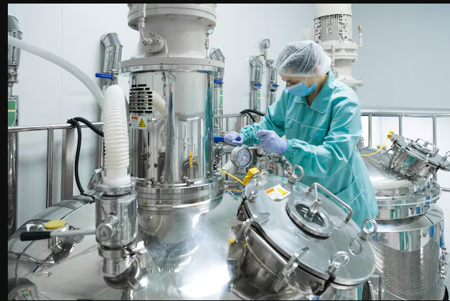 A worker in protective clothing and a hairnet adjusting equipment in a modern chemical processing facility. The worker is inspecting a large, shiny stainless-steel industrial reactor, showcasing the precision and cleanliness required in chemical manufacturing environments.