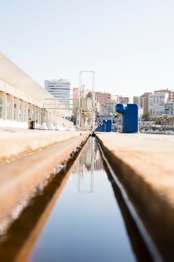 A close-up view of a narrow water channel reflecting buildings and structures on a sunny day. The background shows urban architecture with blue and white industrial equipment.