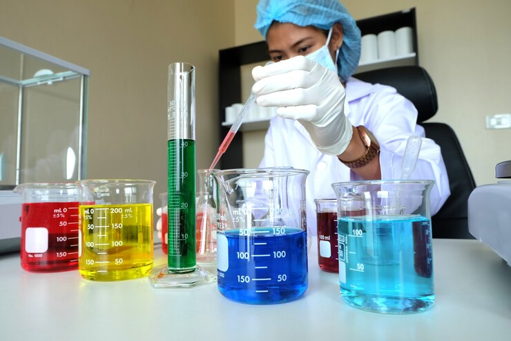 A scientist wearing a lab coat, mask, and hairnet uses a pipette to transfer liquid between beakers filled with colorful solutions. The laboratory setup includes beakers and a graduated cylinder containing red, yellow, green, and blue liquids.