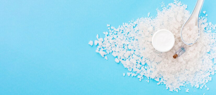 A pile of coarse white crystals spread across a blue background, accompanied by a small white container, a spoon, and a vial, representing salt or a chemical compound used in industrial or culinary applications.