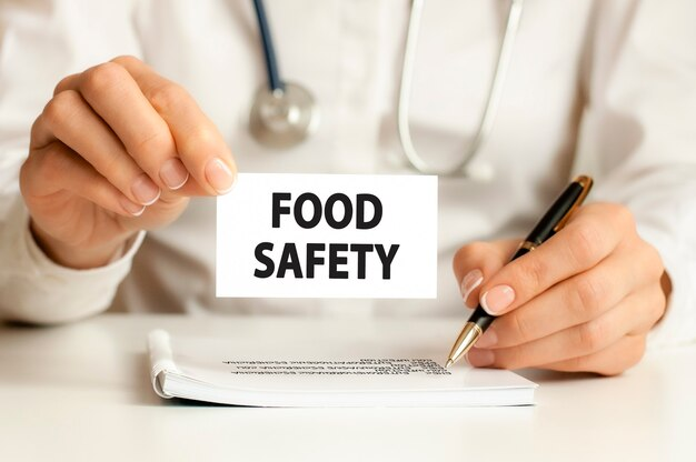 A close-up of a person holding a card with the text 'FOOD SAFETY' while sitting at a desk with a document and pen, wearing a white coat with a stethoscope visible in the background, emphasizing the importance of food safety in healthcare or regulatory contexts