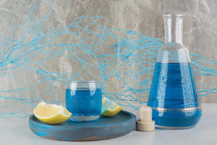 A stylish arrangement featuring a glass carafe and a glass filled with blue liquid on a rustic tray, accompanied by lemon wedges, set against a textured stone background with decorative blue branches.