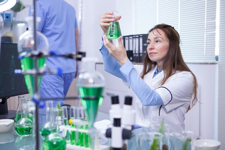 A female scientist in a lab coat examines a flask containing green liquid. The laboratory table is filled with various glassware, including flasks and test tubes, with a focus on experiments involving green solutions.