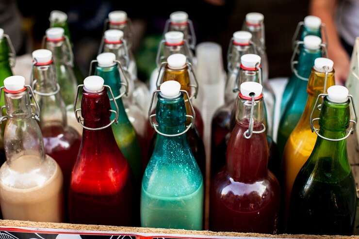 A collection of glass bottles with flip-top caps filled with various colorful liquids, including vibrant red, turquoise, yellow, and brown, displayed at a market or artisanal stall, creating a visually appealing scene