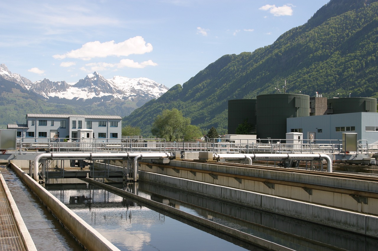 view of a narrow water channel reflecting buildings and structures on a sunny day. The background shows urban architecture with blue and white industrial equipment.