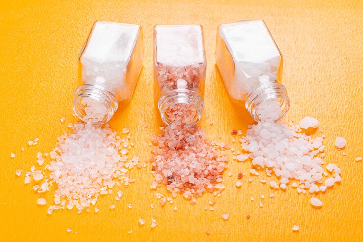 Three glass jars tipped over on a bright orange surface, spilling white and pink coarse crystals, representing different varieties of salts or minerals, showcasing their texture and natural colors.